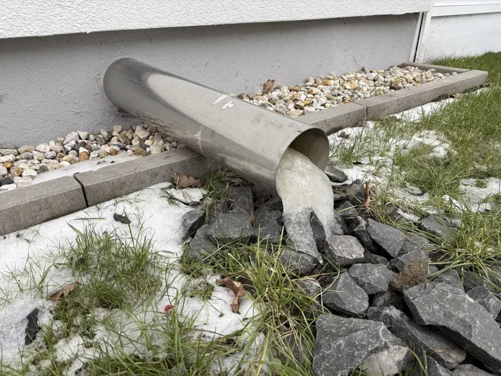 Ice pushing out of the end of a frozen drain pipe beside a house
